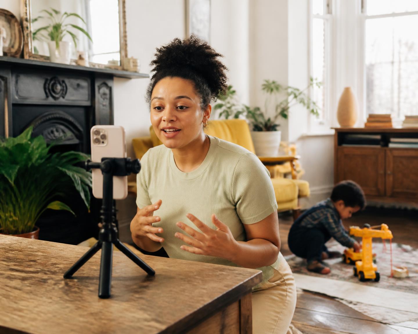 A parent recording content on a phone tripod at home while their child plays nearby