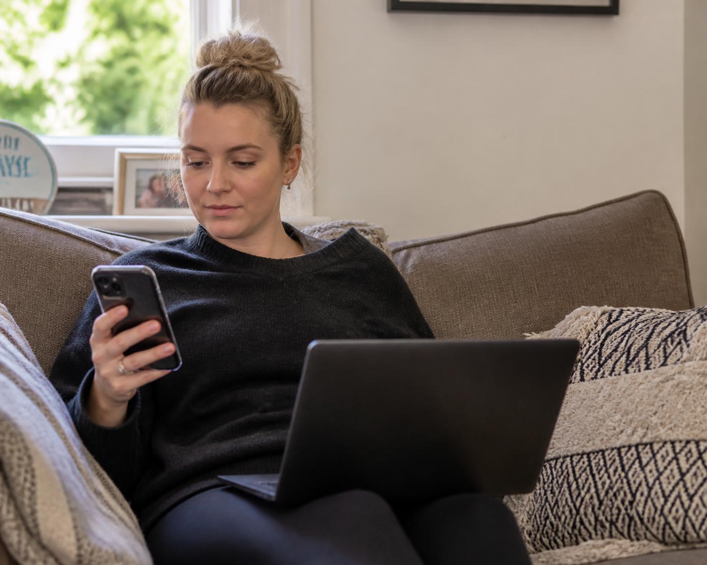A woman sitting on her sofa at home using her phone and laptop