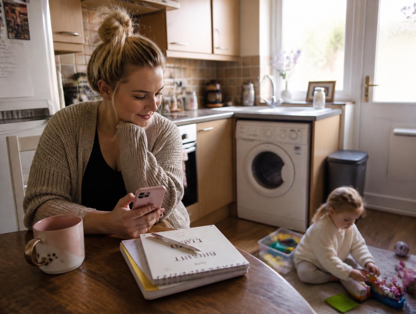 A mother checking her phone at the kitchen table while her toddler plays